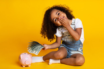 Cute little girl with piggy bank and banknotes, yellow background