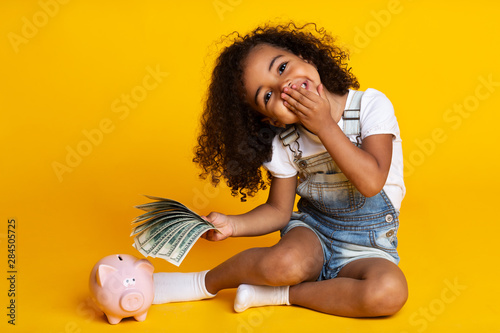 Fotomural Cute little girl with piggy bank and banknotes, yellow background