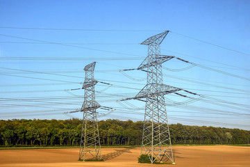 Two pylons of high voltage power lines located on a plowed field, against a background of blue sky and green forest. Shadows from large pillars. Selective focus. Copy space.