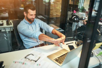 Wall Mural - Relaxing at workplace. Tired young bearded businessman in formal wear stretching his arms and smiling while sitting in the modern office