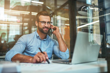 Customer Support. Portrait Of Young And Cheerful Businessman In Eyeglasses And Formal Wear Talking On The Phone And Smiling While Sitting In The Office