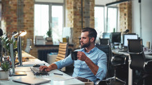 Fresh Coffee. Side View Of Young Bearded Businessman In Formal Wear Holding Cup Of Coffee And Looking At Computer While Sitting In The Modern Office