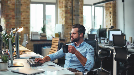 Fresh coffee. Side view of young bearded businessman in formal wear holding cup of coffee and looking at computer while sitting in the modern office