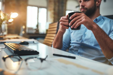 Coffee break. Young bearded businessman in formal wear drinking coffee and looking at computer while sitting in the modern office