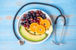 © Jo Panuwat D - Top view black grapes, red grapes, Valencia orange and melon fruits in white dish with Cardiology Stethoscope on blue wood table. dieting healthy food and healthcare concept