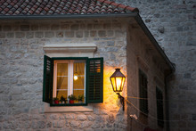 Window With Wooden Shutters And Street Light On Stone Wall. Ancient Authentic House With Tiled Roof In Old Town At Night. Windowsill Is Decorated With Red Flowers. Kotor, Montenegro