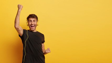 Attractive young man feels happy and overjoyed, lifts clenched fists, enjoys good song vibes, wears headphones, dances actively, isolated on yellow background with free space for your promotion