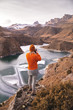 Portrait from the back of the girl traveler photographer in an orange sweater and hat with a camera in hand in the mountains against the background of a frozen mountain lake. Photo travel concept