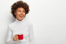 Joyful Pleased Dark Skinned Female Drinks Tea From Red Cup, Looks On Right Side, Glad To Have Spare Time, Enjoys Coffee Break, Dressed In Casual Outfit, Isolated On White Studio Wall, Copy Space
