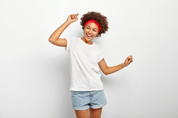 Wall Mural - Carefree pleased teenage girl has fun, dances joyfully with raised arms, being entertained and amused, wears summer clothes, laughs happily, enjoys cool music, isolated on white wall, being on party