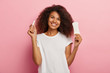 Isolated shot of happy young Afro female holds menstuation cotton tampon and sanitary napkin, demonstrates intimate products for women hygiene, wears casual white t shirt, has dark curly hair