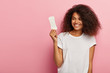 Satisfied lovely woman with crisp hairstyle, holds sanitary napkin, uses hygienic product during critical days, smiles positively, wears mockup t shirt, isolated on pink wall. Women health concept