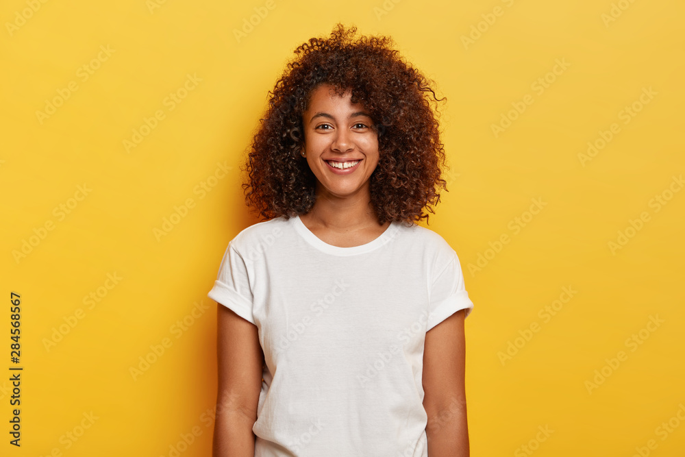 Headshot of beautiful dark skinned curly has pleased expression, rejoices success, enjoys spare time, wears casual t shirt, isolated on yellow background. People, positive emotions, feelings concept - obrazy, fototapety, plakaty 