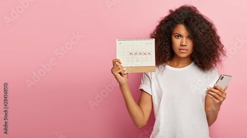 Cuadros en Lienzo Displeased dark skinned woman has unhappy look, holds periods calendar with mark