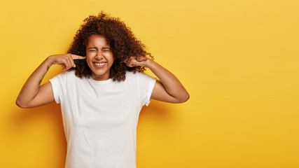 Studio shot of lovely stressed African American woman clenches teeth, doesnt want to listen anything, hears heartbreaking scream, plugs ears with index fingers, wears white t shirt, isolated on yellow