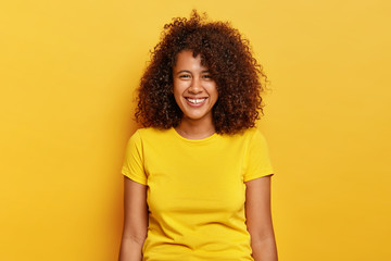 Lovely young female with crisp hair has fun during weekend, tender smile, wears casual yellow t shirt, enjoys summer, gazes satisfied, stands indoor. Monochrome shot. Emotions and feelings concept