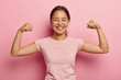 © WHstudio Leushin N - Strong powerful Asian woman with dark combed hair, toothy smile, raises arms and shows biceps, has piercing in ear, wears casual rosy t shirt, models against pink background. Look at my muscles!