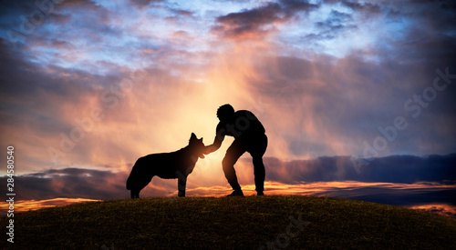 Cuadros en Lienzo Silhouette of a man caressing his dog on a hill at sunset.