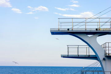 White diving board or tower against a clear blue sky. In the background the sea.