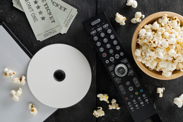 DVD or blu ray movie disc with tv remote control, movie tickets and bowl of popcorn on dark background. Home theatre movie or series night concept. Flat lay from above.