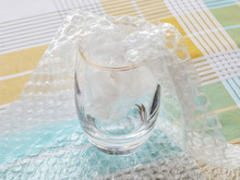 Thick Bottom Glass Half Packaged With Transparent Bubble Wrap On A Checkered Tablecloth. Material For Packing Fragile Items For Safe Transportation.