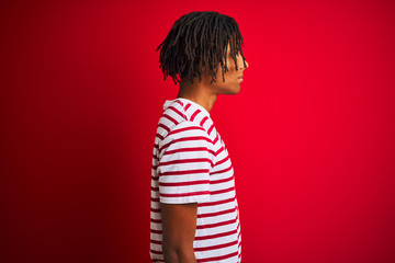 Wall Mural - Young afro man with dreadlocks wearing striped t-shirt standing over isolated red background looking to side, relax profile pose with natural face and confident smile.