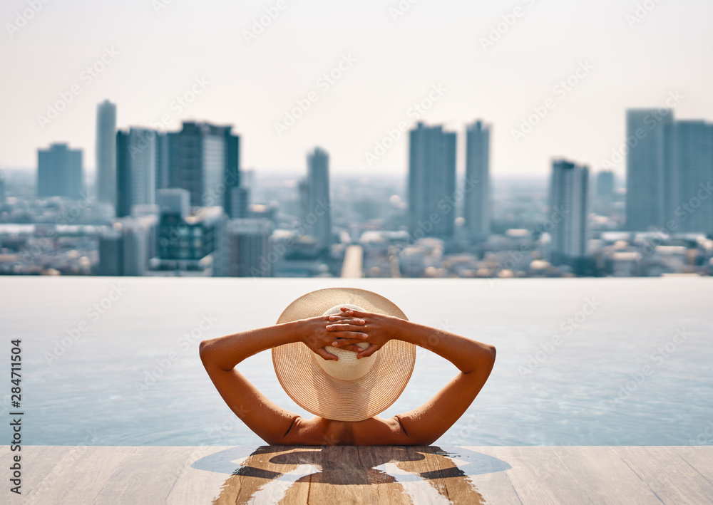 Back view of young woman in hat relaxing in swimming pool on the roof ...
