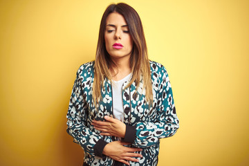 Sticker - Young beautiful woman wearing casual jacket over yellow isolated background with hand on stomach because indigestion, painful illness feeling unwell. Ache concept.