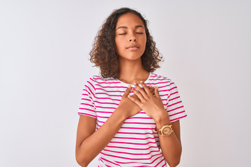 Wall Mural - Young brazilian woman wearing pink striped t-shirt standing over isolated white background smiling with hands on chest with closed eyes and grateful gesture on face. Health concept.