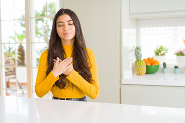 Poster - Young beautiful woman at home on white table smiling with hands on chest with closed eyes and grateful gesture on face. Health concept.