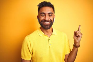 Young indian man listening to music using earphones standing over isolated yellow background with a big smile on face, pointing with hand and finger to the side looking at the camera.