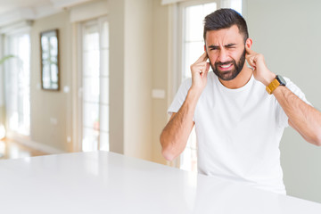 Poster - Handsome hispanic man casual white t-shirt at home covering ears with fingers with annoyed expression for the noise of loud music. Deaf concept.