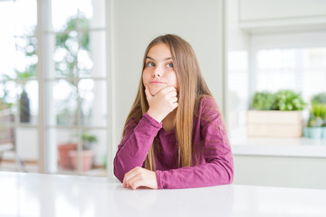 Beautiful young girl kid on white table looking confident at the camera with smile with crossed arms and hand raised on chin. Thinking positive.