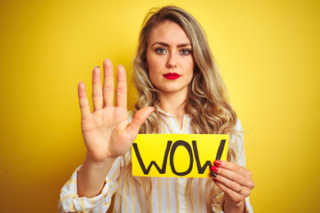 Sticker - Beautiful woman holding amazed wow surprise banner over isolated yellow background with open hand doing stop sign with serious and confident expression, defense gesture