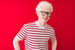 © Krakenimages.com - Young albino blond man wearing striped t-shirt and glasses over isolated red background winking looking at the camera with sexy expression, cheerful and happy face.