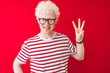 © Krakenimages.com - Young albino blond man wearing striped t-shirt and glasses over isolated red background showing and pointing up with fingers number three while smiling confident and happy.