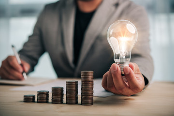 Poster - businessman hand holding lightbulb with coins stacking on desk.saving energy and money concept.