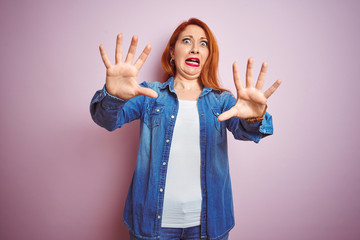 Wall Mural - Youg beautiful redhead woman wearing denim shirt standing over isolated pink background afraid and terrified with fear expression stop gesture with hands, shouting in shock. Panic concept.