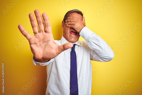 Fotomural Handsome middle age businessman standing over isolated yellow background covering eyes with hands and doing stop gesture with sad and fear expression