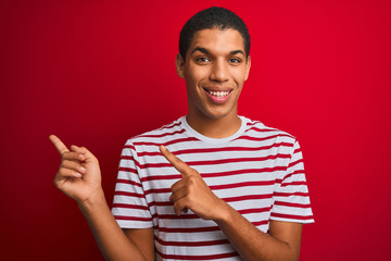 Young handsome arab man wearing striped t-shirt over isolated red background smiling and looking at the camera pointing with two hands and fingers to the side.