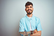 © Krakenimages.com - Young man with tattoo wearing blue shirt standing over isolated white background happy face smiling with crossed arms looking at the camera. Positive person.