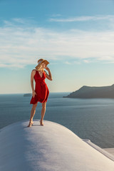 Wall Mural - blond woman in a red dress and a sun hat, in Oia on Santorini, standing on a roof, with the caldera and aegean sea in the background