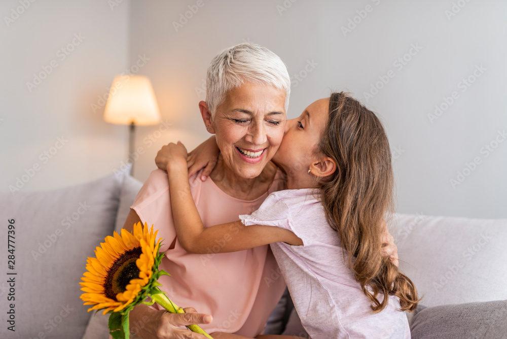 Happy grandmother hugging small cute grandchild thanking for flowers presented, excited granny embrace granddaughter congratulating her with birthday, making surprise presenting bouquet - obrazy, fototapety, plakaty 