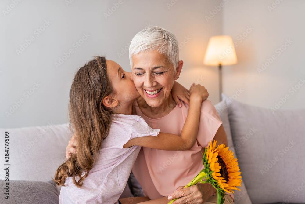 Happy senior grandma hugging granddaughter thanking for gift and flowers. Little granddaughter kissing giving flowers bouquet congratulating smiling old grandmother - obrazy, fototapety, plakaty 