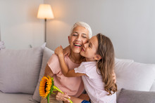  Happy Senior Grandma Hugging Granddaughter Thanking For Gift And Flowers. Little Granddaughter Kissing Giving Flowers Bouquet Congratulating Smiling Old Grandmother