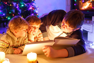 Father and two little toddler boys reading book by chimney, candles and fireplace.