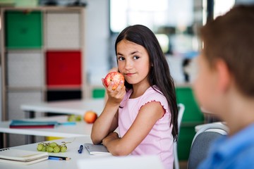 Wall Mural - A small school girl sitting at the desk in classroom, eating apple.