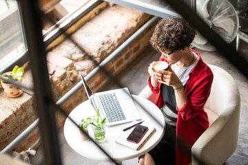 Wall Mural - Young woman with cup of cappuccino sitting by table in cafe in front of laptop