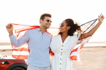 Wall Mural - Photo of joyous multiethnic couple smiling and holding american flag while standing by car outdoors