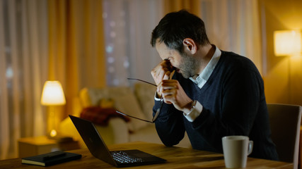 Aufkleber - Tired Middle Aged Man Works on a Laptop. He's Eyes Fatigued, He Temporarily Takes off His Glasses and Rubs His Eyes. His Apartment Looks cosy in Yellow Tones.
